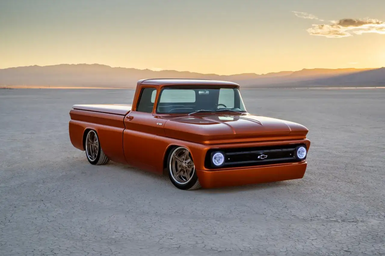 Pomarańczowy Chevrolet pickup, klasyczne modele z lat 60., lśniące felgi, pustynny krajobraz o zachodzie słońca.