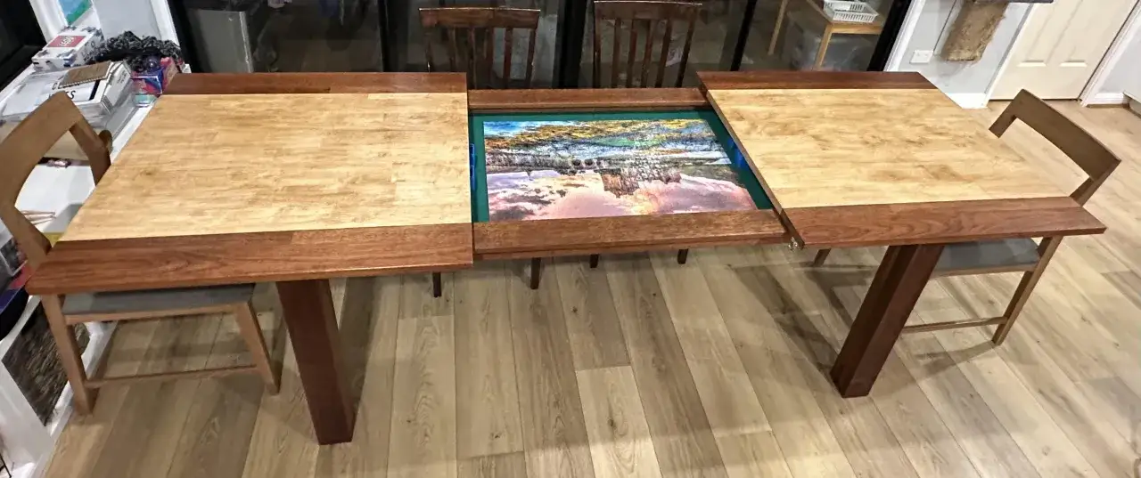 St&oacute;ł z wysuwanymi blatami, idealny do układania puzzli. Widoczny fragment obrazu z mostem i niebem.